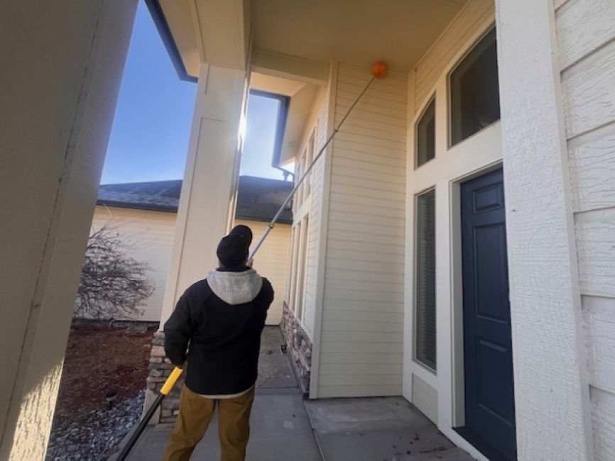 A person uses a long, extendable pole to reach and clean a high corner on the exterior of a cream-colored house.