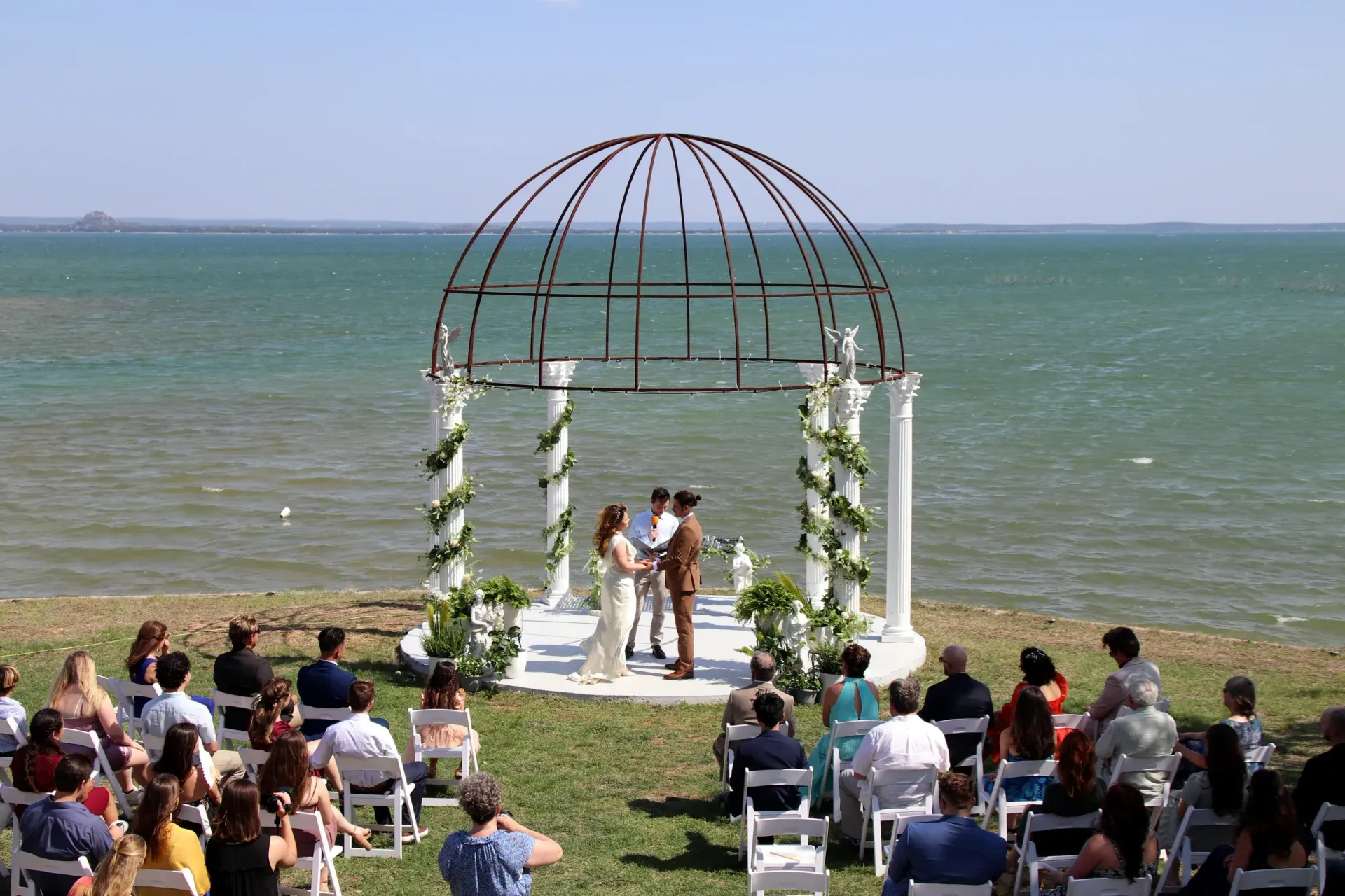 Wedding ceremony under a white arch by the water, with guests seated facing the couple on a grassy shore.