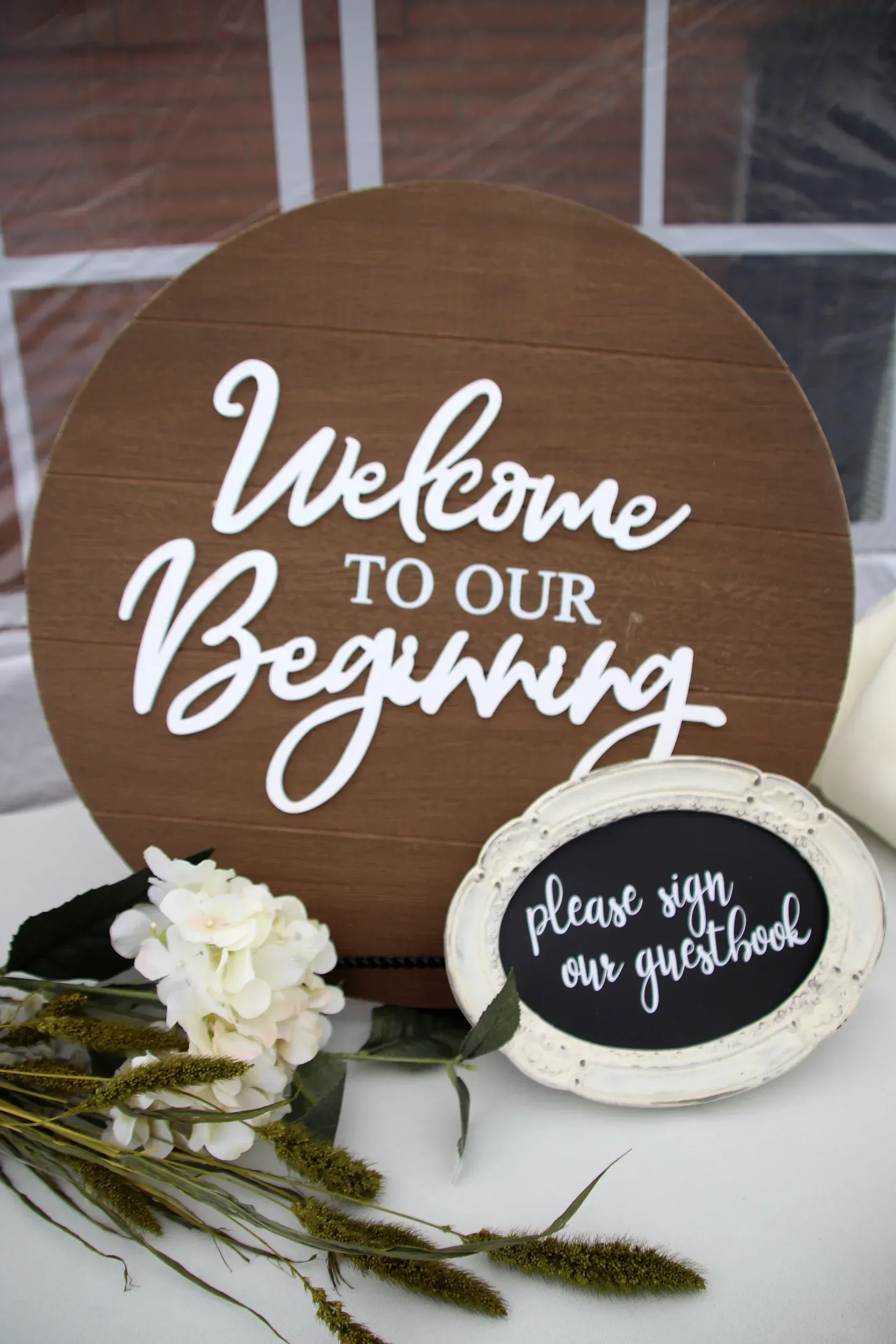 Brown wedding sign reading “Welcome to Our Beginning” with white flowers and a small “please sign our guest book” plaque