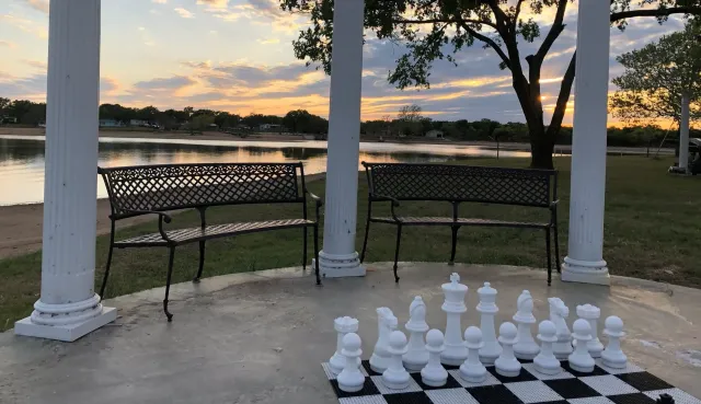 Outdoor chess set beside a lakeside gazebo at sunset with white columns and benches