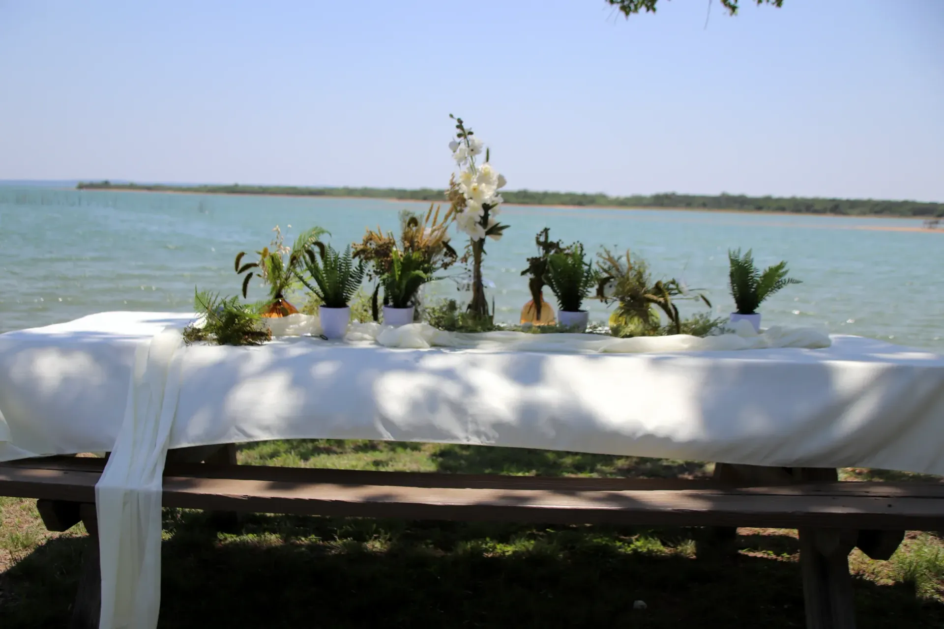 White-clothed table with flowers beside a calm lake under a clear blue sky