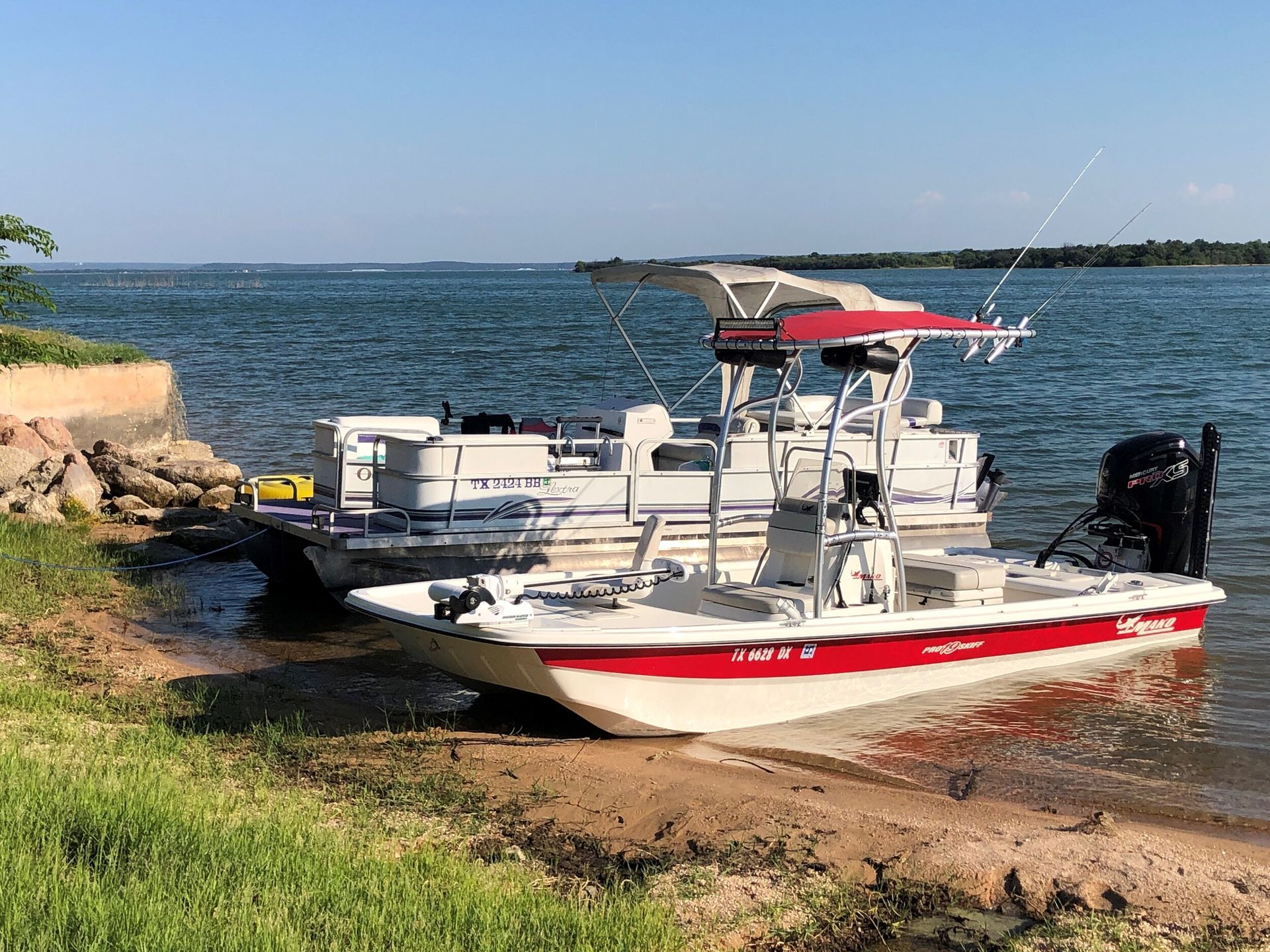Small red-and-white motorboat on a sandy shore by calm blue water, with a dock and green grass nearby