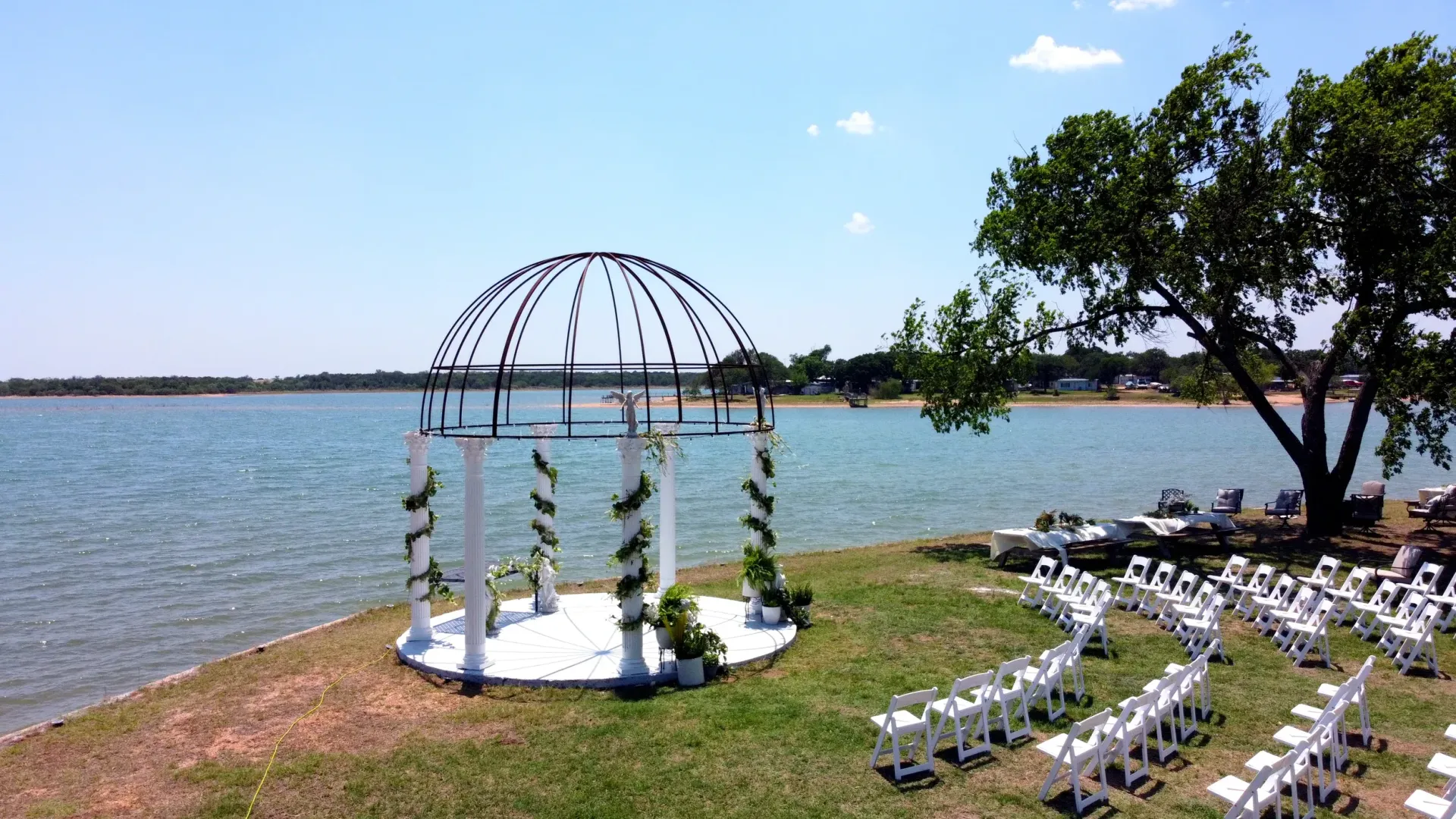 Lakeside wedding setup with white chairs, floral arch, and water view under a blue sky