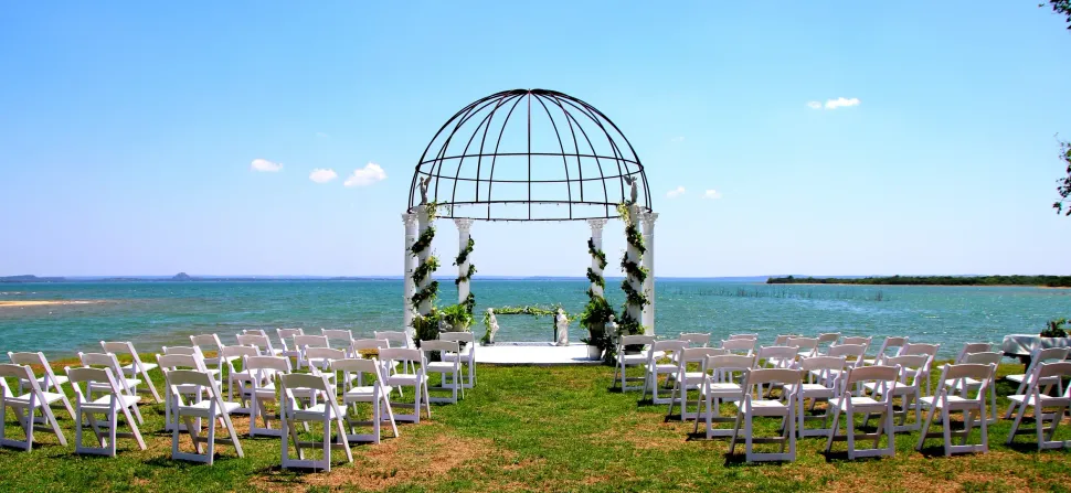 Outdoor wedding ceremony setup by the water with white chairs and a floral arch under a blue sky