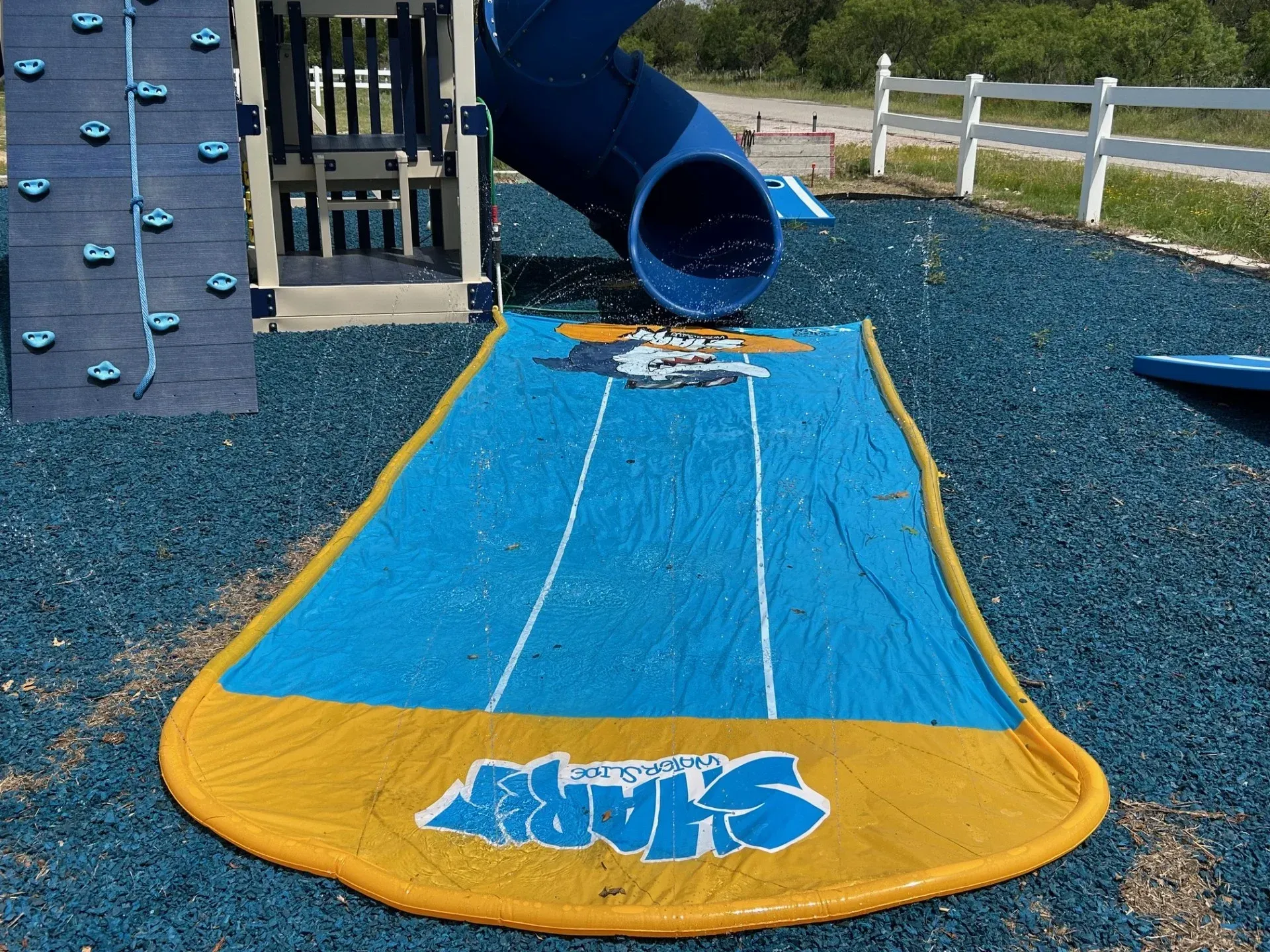 Blue playground slide with a yellow landing mat on a mulch surface, next to climbing equipment and a fence
