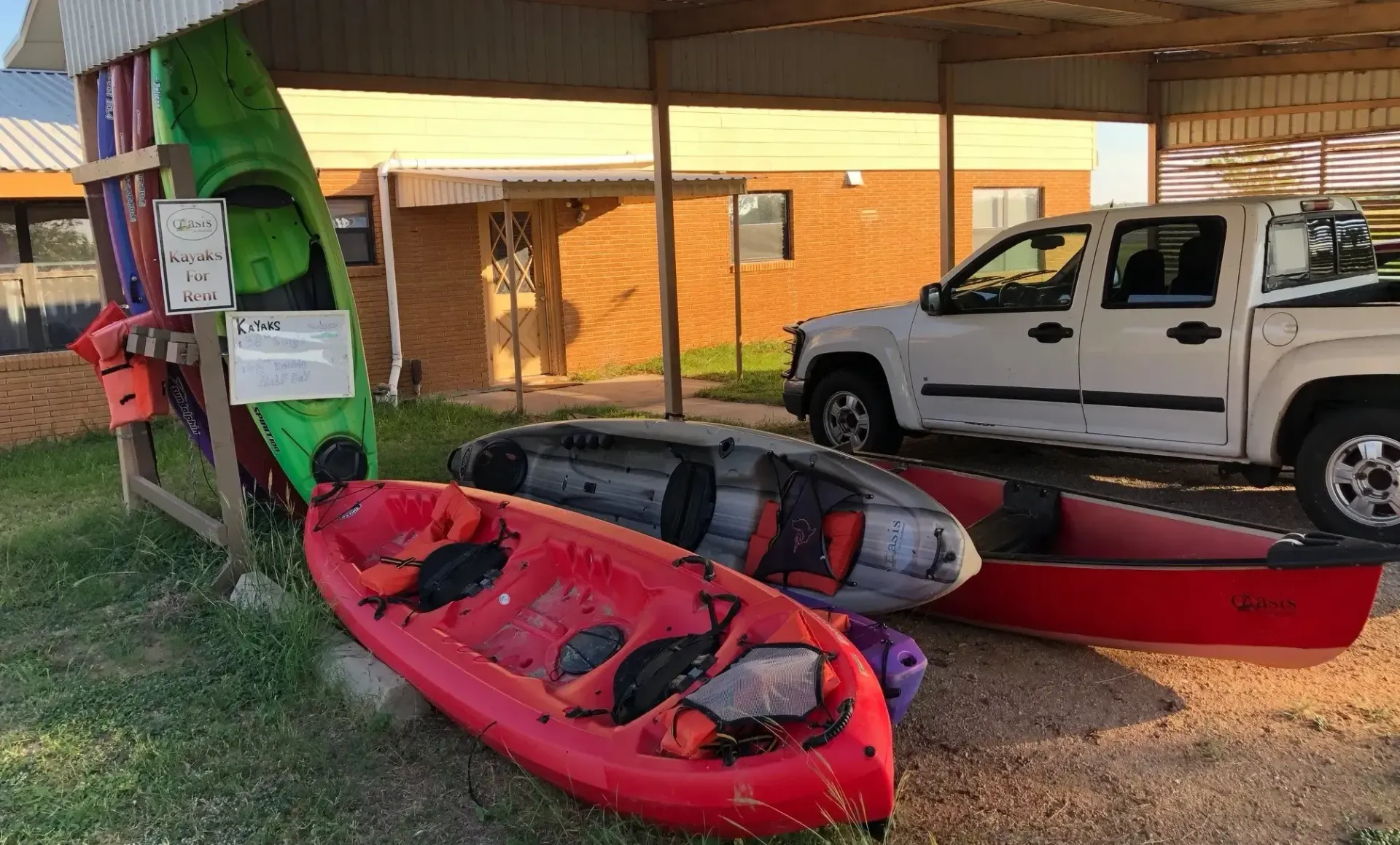 Red kayak, two black kayaks, and a white truck parked under a canopy beside a brick building.