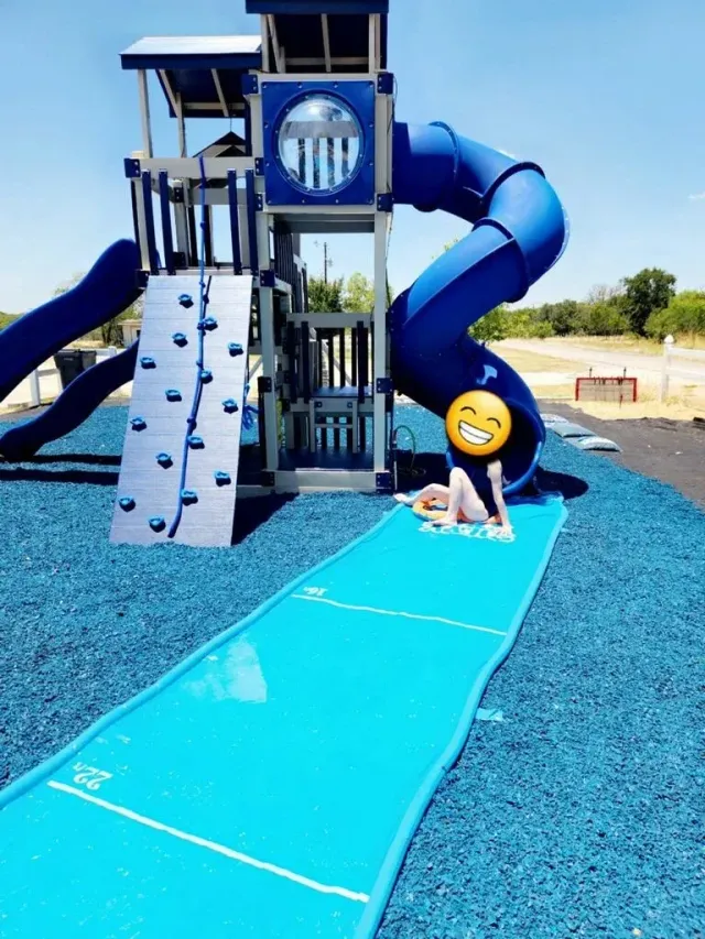 Child on a playground slide beside blue climbing equipment and a turquoise path under a bright sky