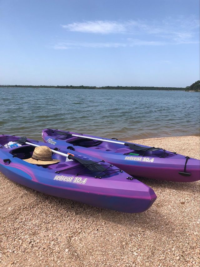 Two purple kayaks on a pebbled beach beside calm water under a blue sky