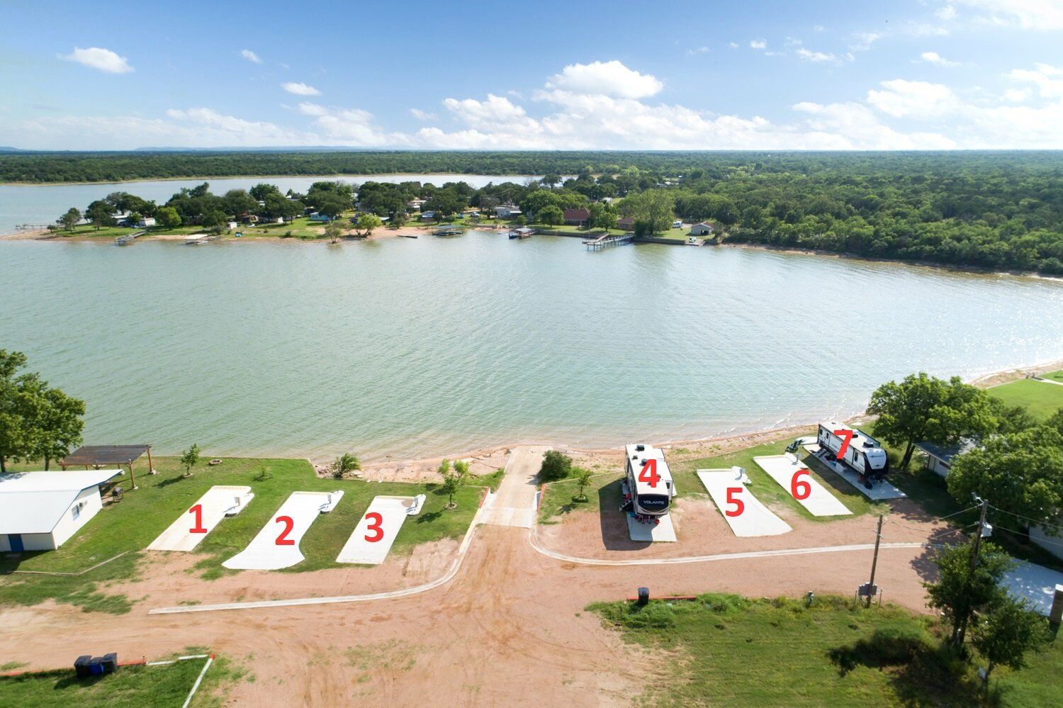Lakeside sandy lot with numbered concrete pads and a small boat near the water under a clear sky