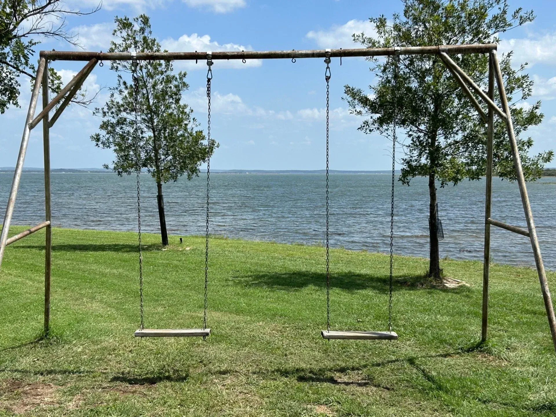 Wooden swing set on a grassy lakeshore with trees and blue water in the background