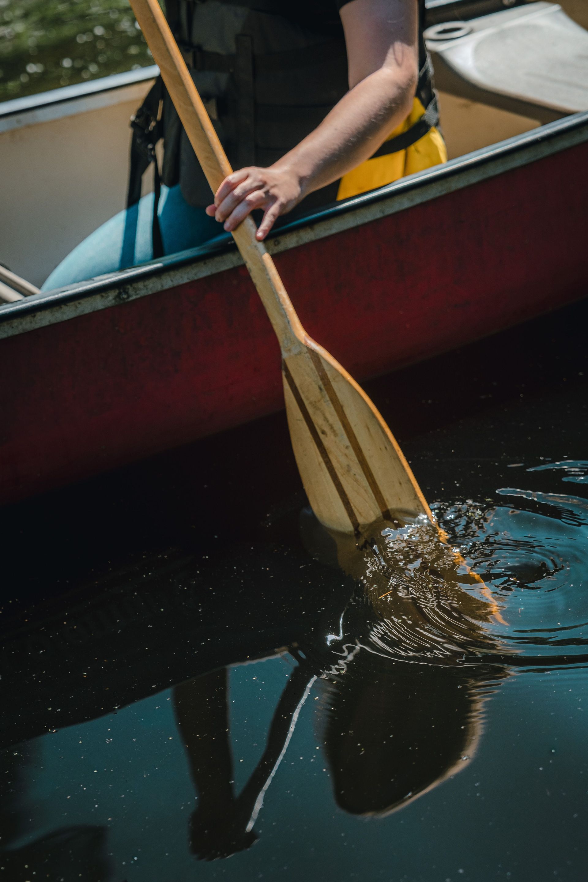 Person paddling a red canoe on dark water, with a wooden oar dipping into the lake.