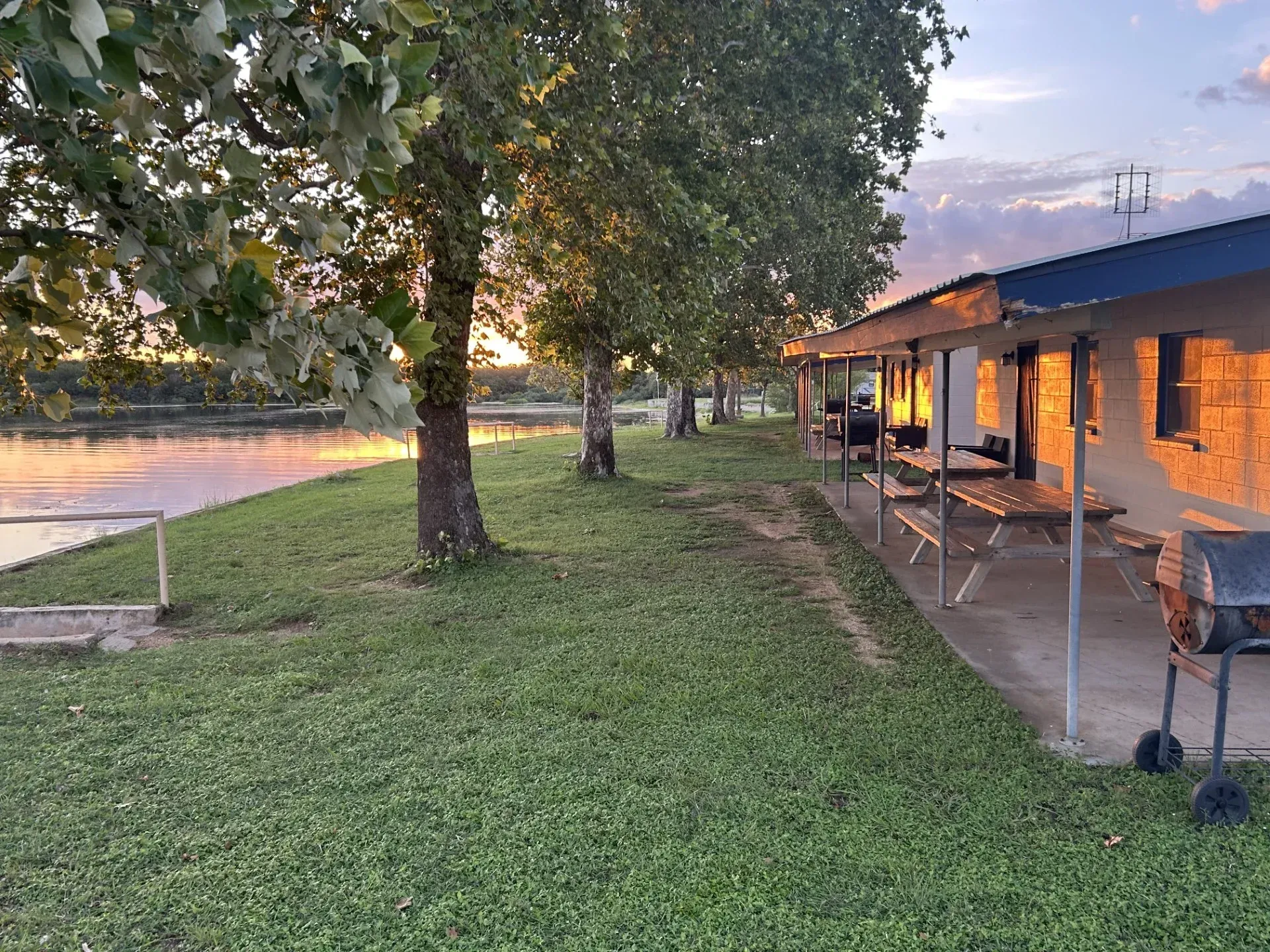 Sunset over a riverside lawn beside a lit porch and house with trees along the water