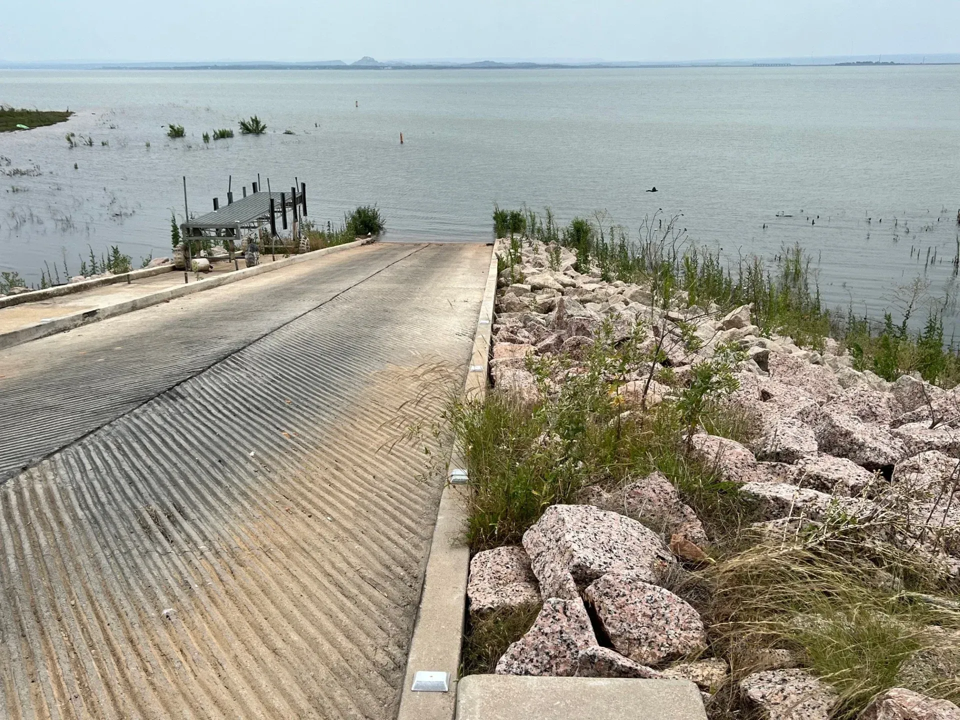 Concrete boat ramp leading into a calm lake beside rocky shoreline and sparse grass