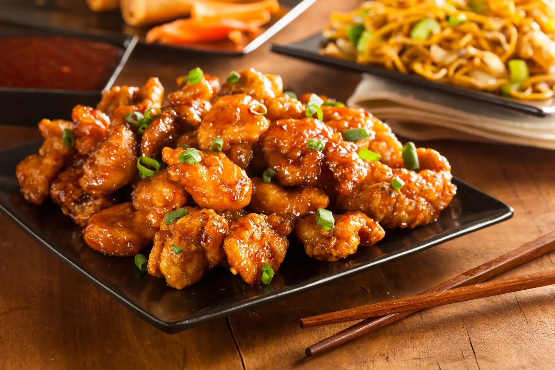 Glazed fried chicken bites on a black plate with chopsticks on a wooden table, with side dishes in the background