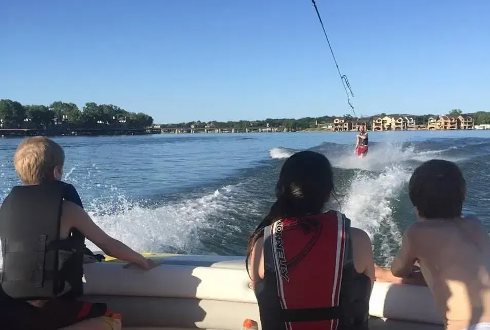 Three people ride on a boat, watching another boat speed across a sunny lake.