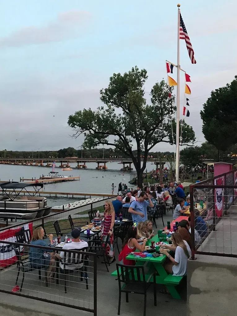 Outdoor waterfront dining area with people seated at tables beside a dock and flagpoles at sunset