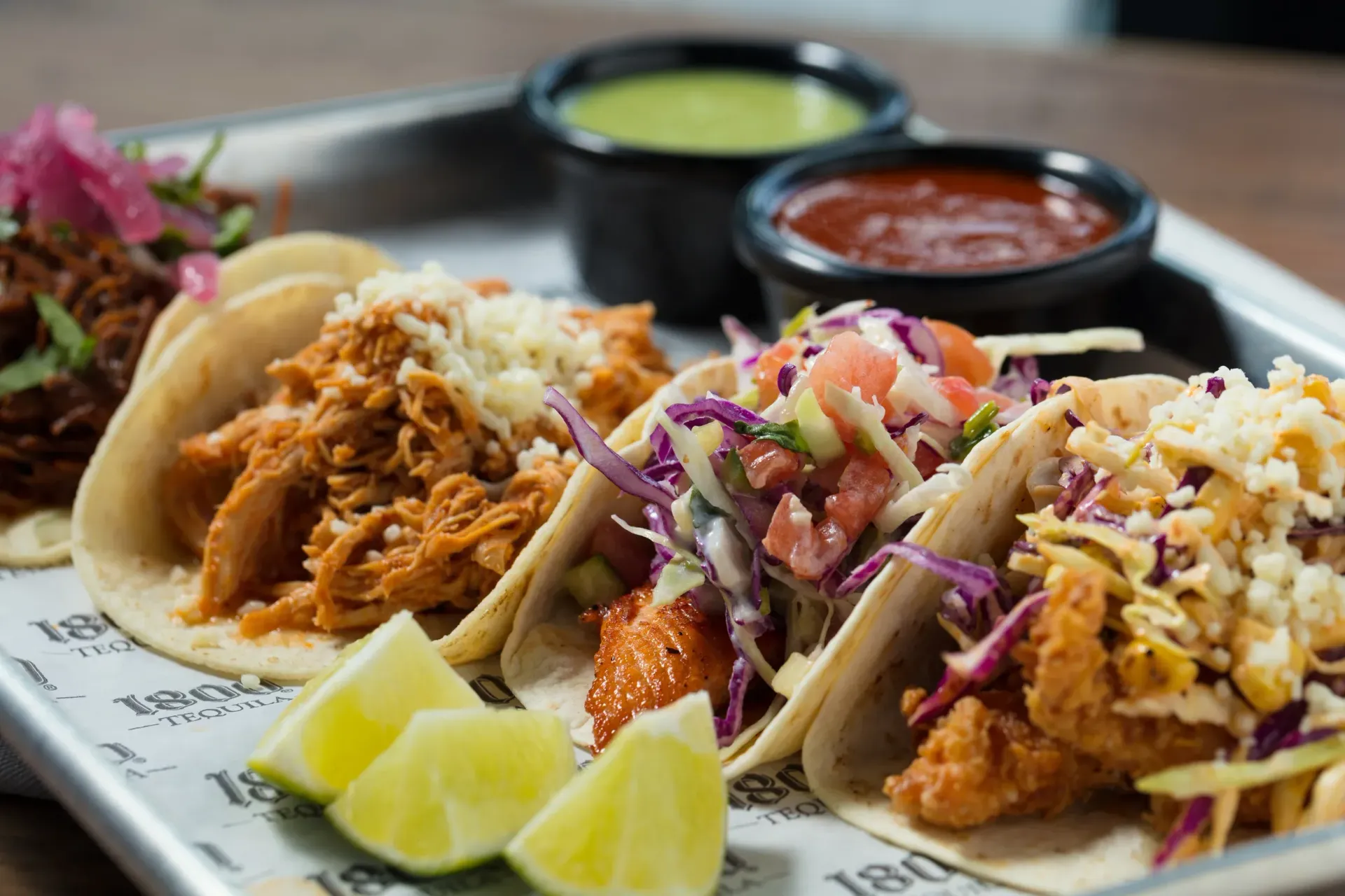 Tacos with shredded meat, slaw, and salsa on a tray with lime wedges and dipping sauces