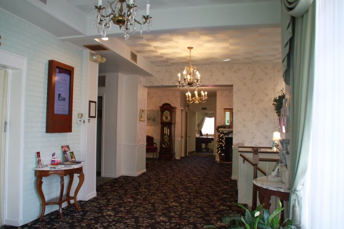 Hallway with floral carpet, chandeliers, grandfather clock, and a table with display items.