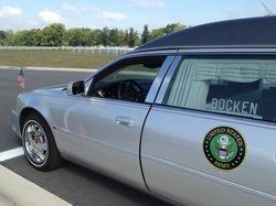 Silver hearse with US Army emblem, parked on pavement. 