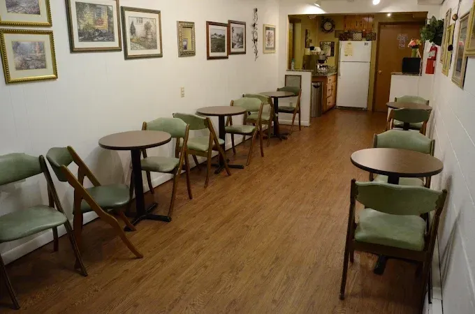 A small, sparsely furnished room with round tables, green chairs, and framed pictures on the walls.