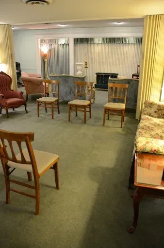 A funeral home parlor with chairs arranged on a green carpet, beige curtains, and dim lighting.