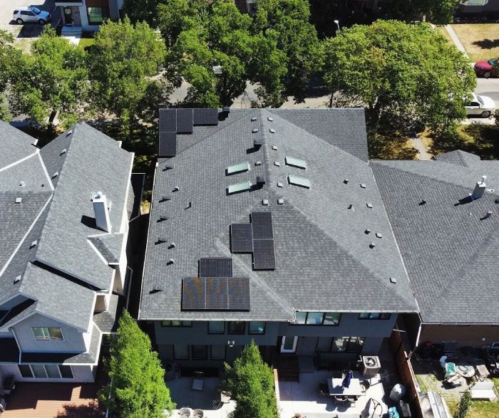 Aerial view of a house with solar panels on a dark gray roof, surrounded by trees and neighboring homes.