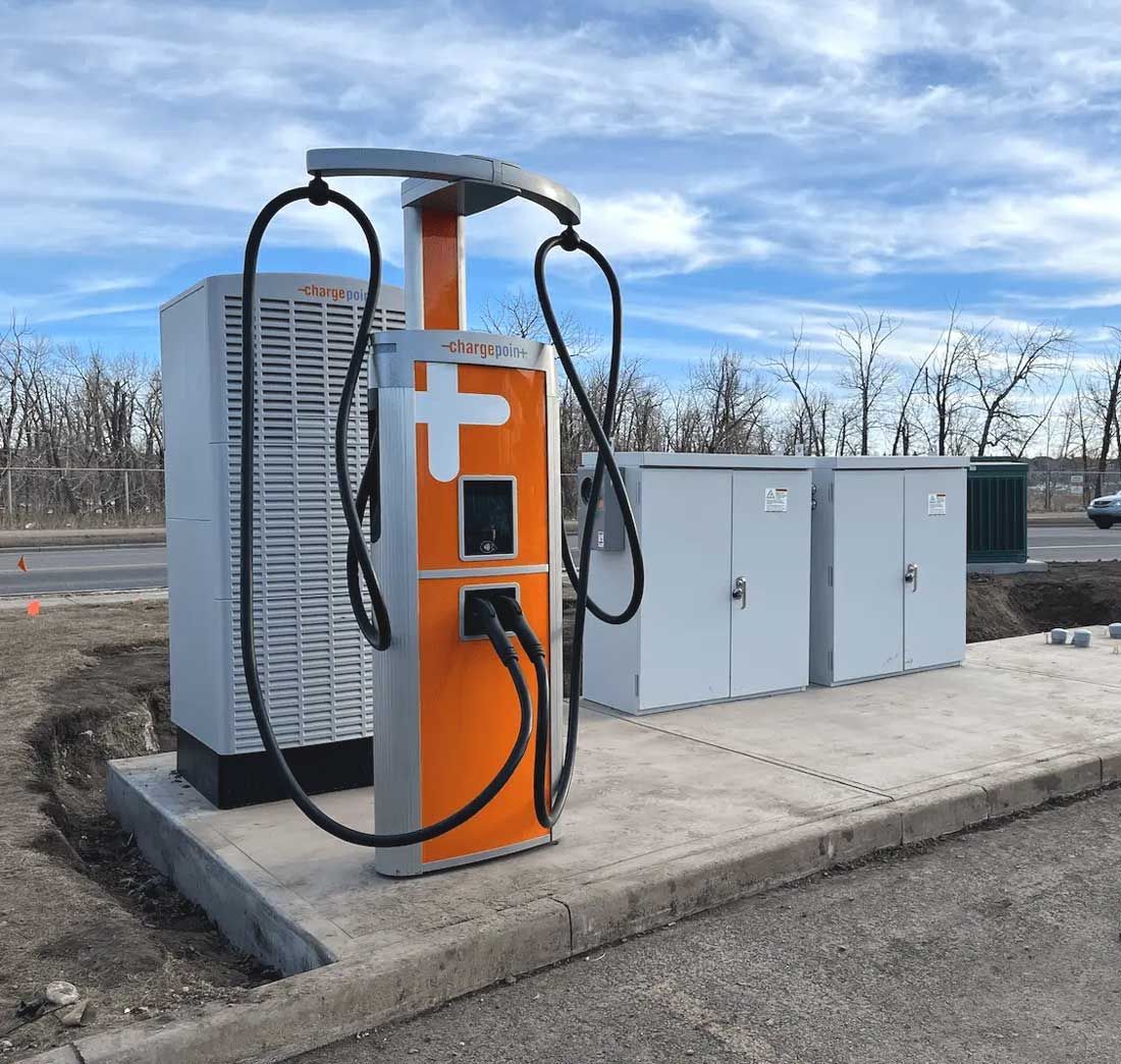 Electric vehicle charging station with orange and silver accents; cables hanging; cloudy sky in background.