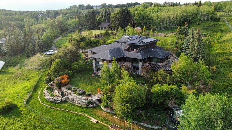 Large stone house with dark roof surrounded by green trees on a hillside.