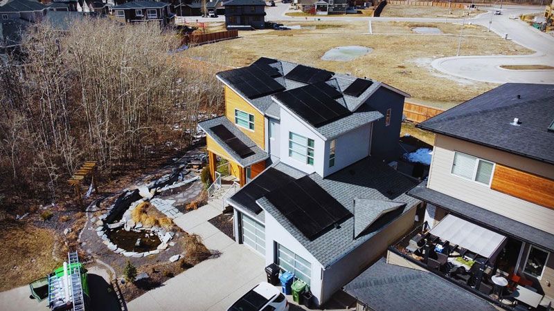 House with solar panels on roof, front yard with landscaping. Houses in the background. Sunny day.