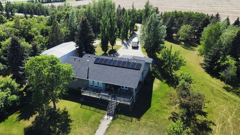 Aerial view of a house with solar panels on a dark roof, surrounded by trees and green grass. A vehicle is on the driveway.