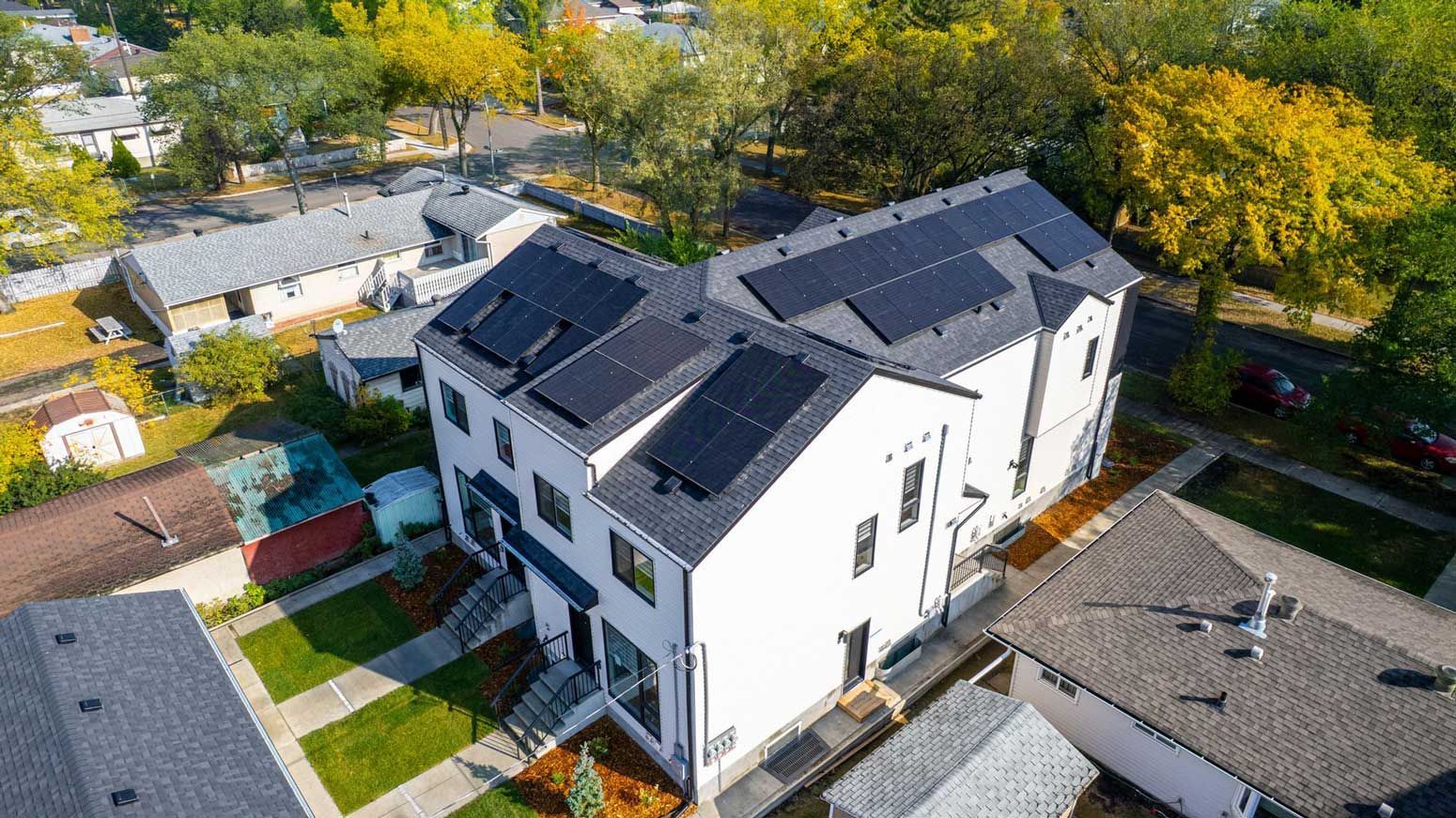Aerial view of a house with solar panels on a dark roof, surrounded by trees and green grass. A vehicle is on the driveway.