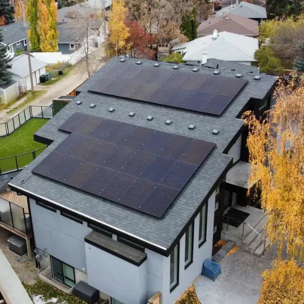 Aerial view of a house with solar panels on the dark gray roof, surrounded by other houses and trees with fall foliage.