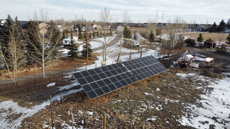 Solar panels in a snowy field, angled towards the sky. Trees and houses are in the background.