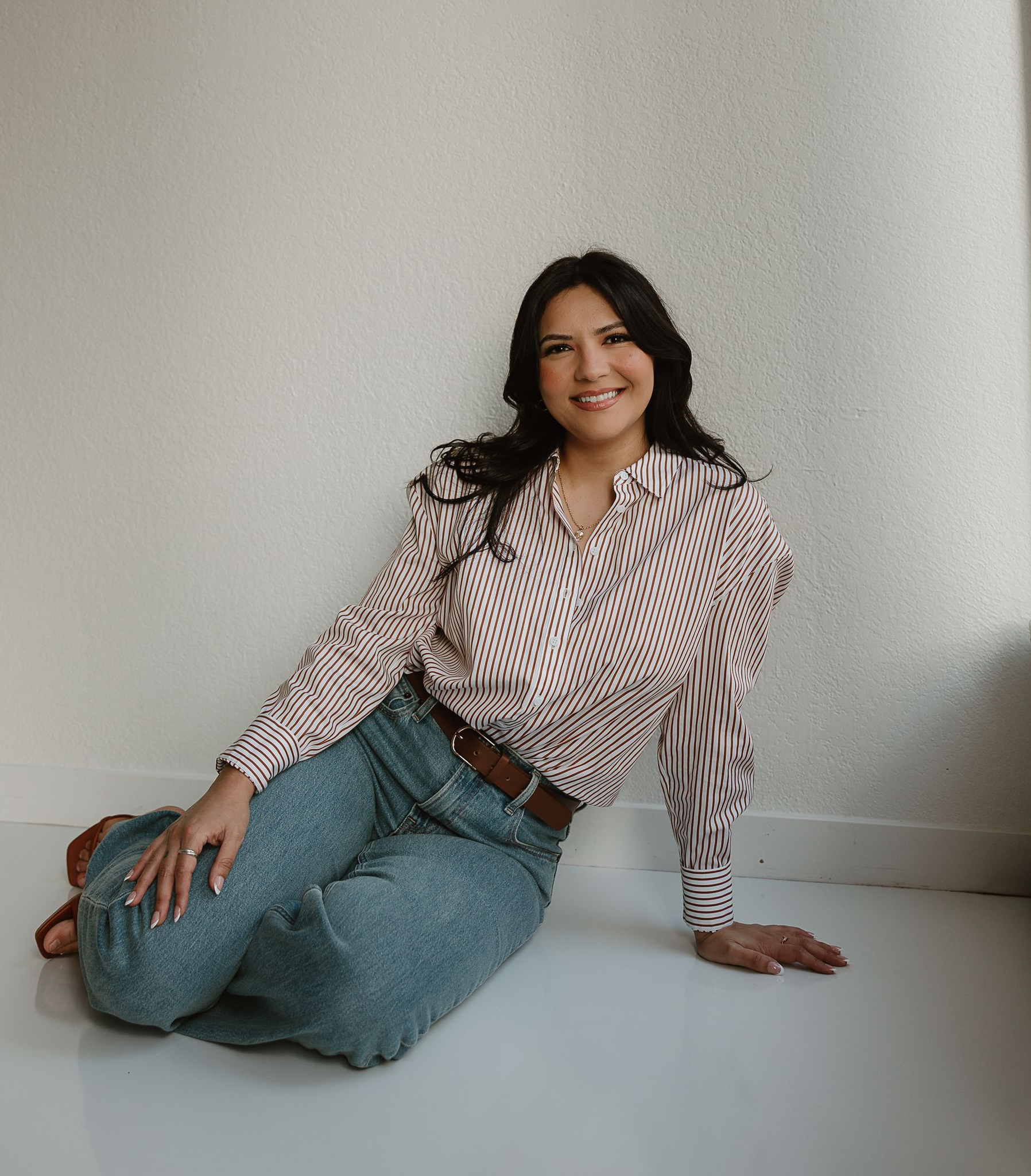 A smiling person with long, dark wavy hair sits on a white floor against a white wall, wearing a patterned shirt and jeans.