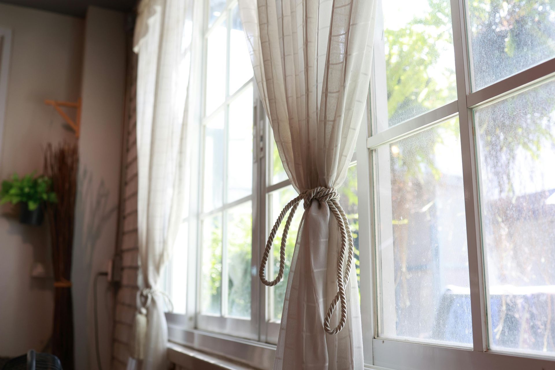 Light, airy curtains gathered with decorative rope ties hang in front of a white-framed window looking out to a garden.