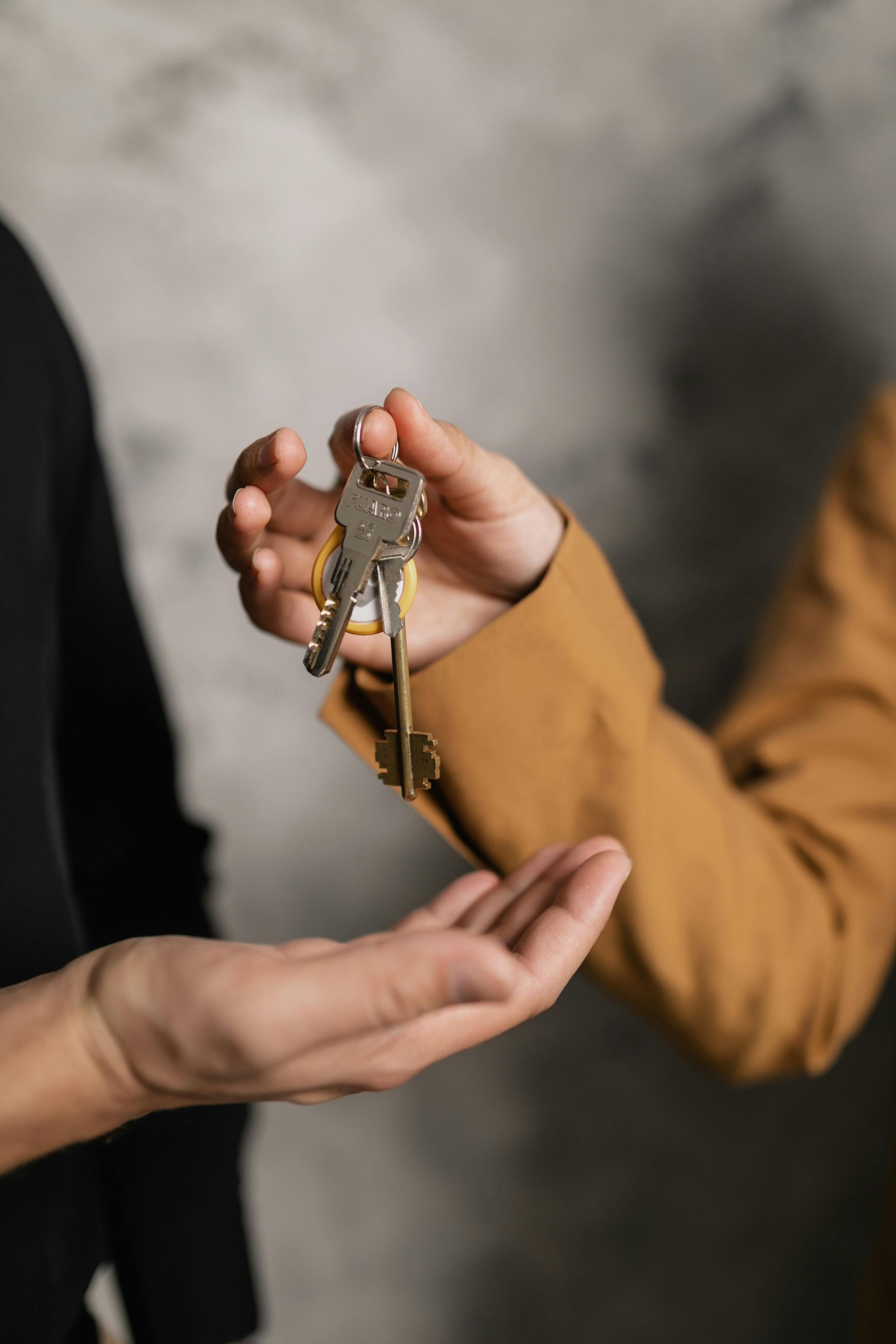 Hands exchanging keys, one wearing a tan blazer, the other black, against a textured gray background.
