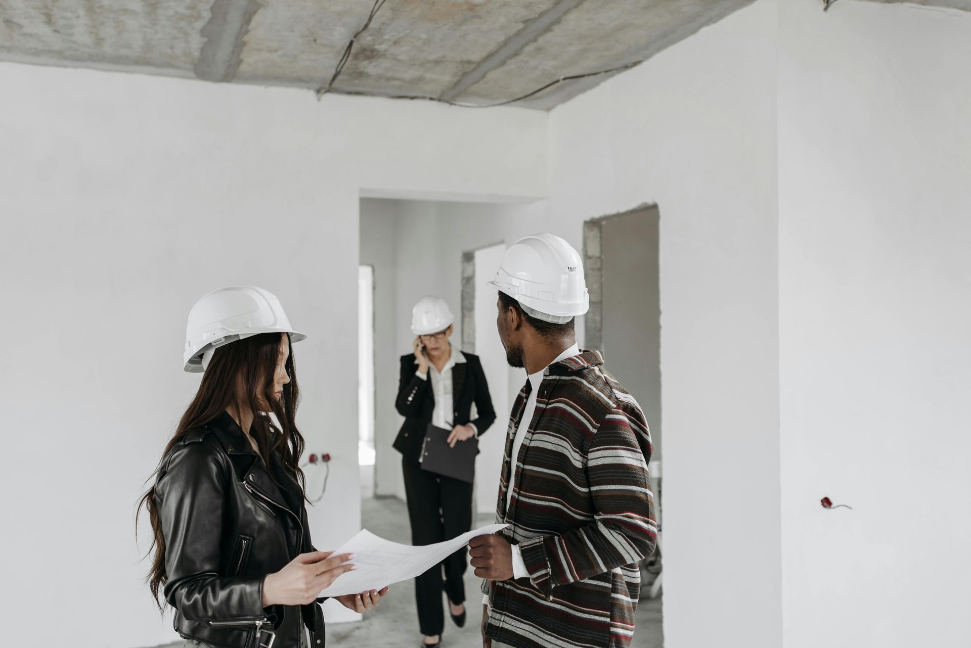 Three people in hard hats inside a building under construction, reviewing blueprints.