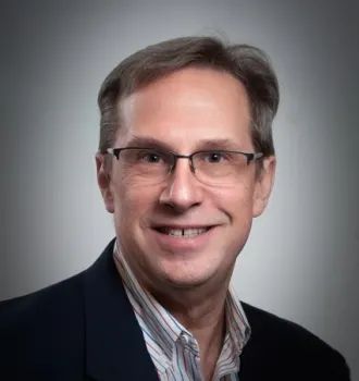 Man with glasses smiles, wearing a striped shirt and navy blazer against a gray backdrop.