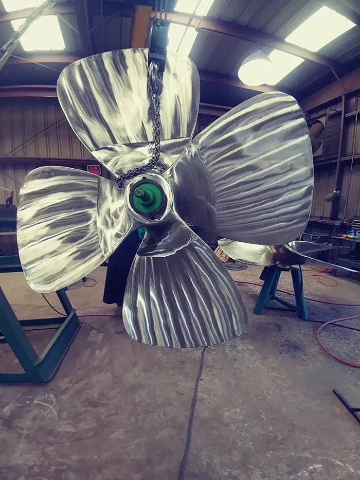 A large stainless steel propeller is hanging from a chain in a warehouse.