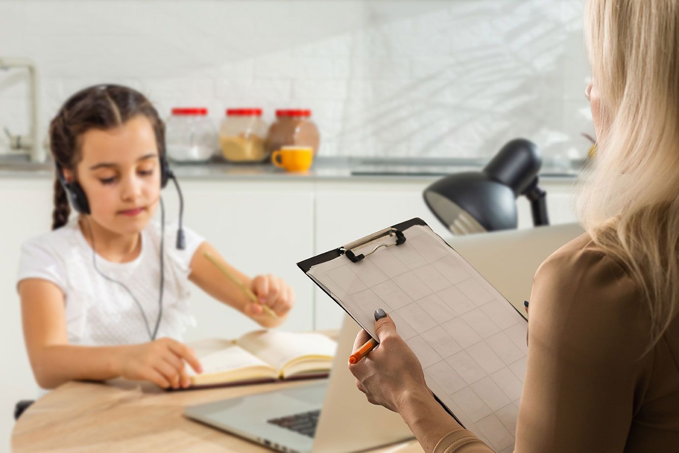 Tutor observing a child writing at a kitchen table while holding a clipboard and pen