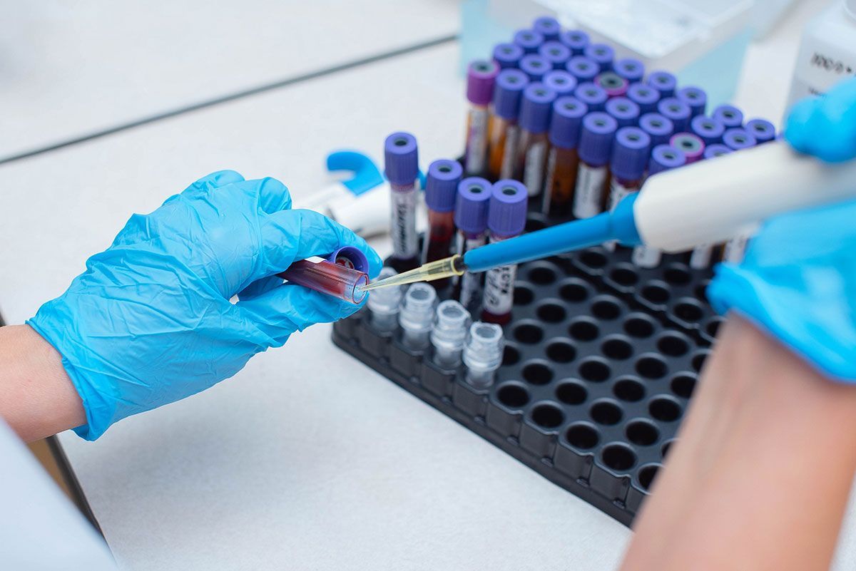 Gloved hands using a pipette to transfer liquid into test tubes in a lab rack