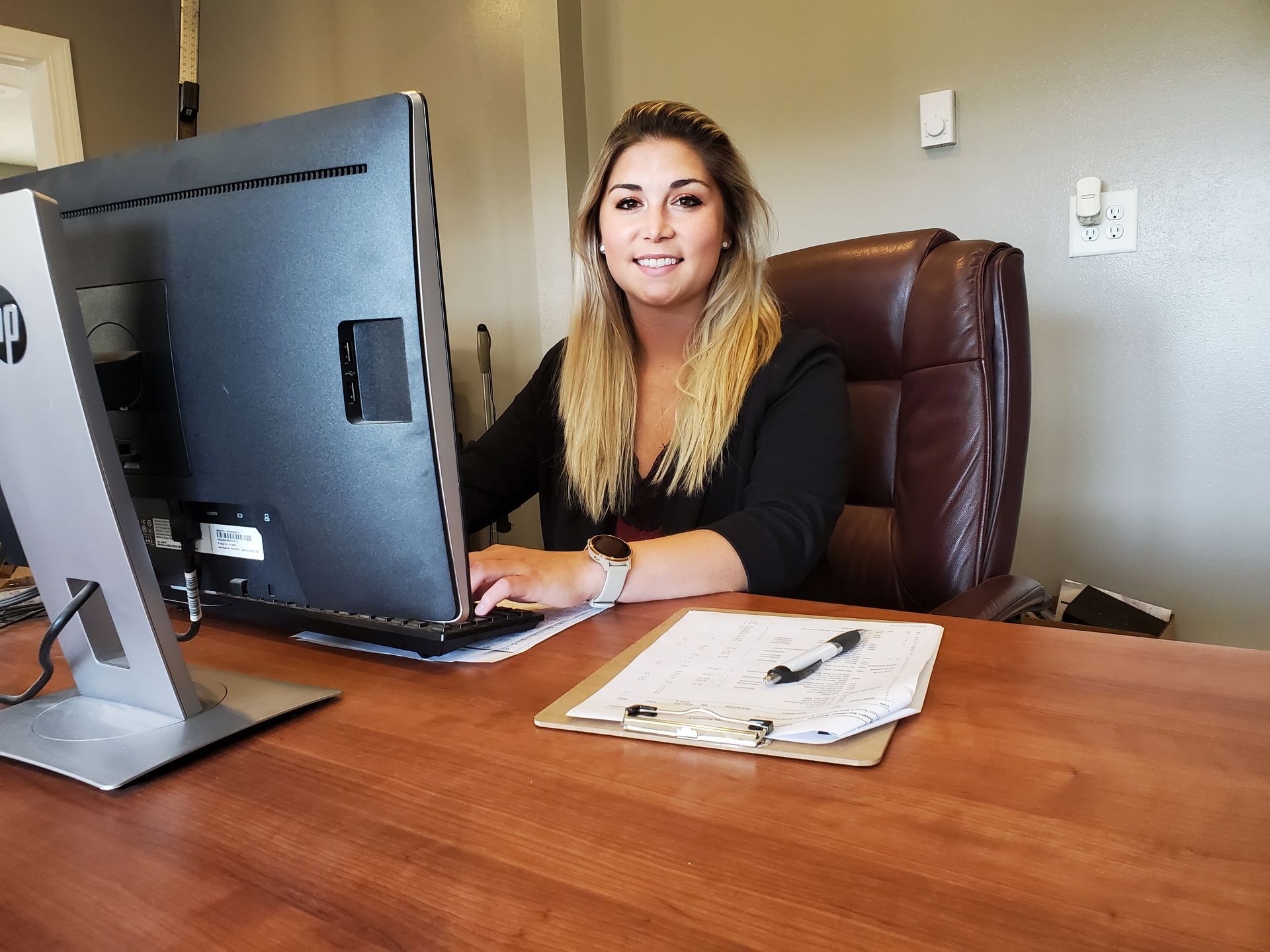 Woman seated at a desk in an office, smiling beside a computer and clipboard