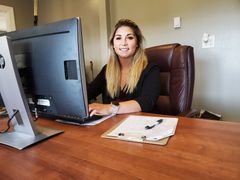 Woman seated at a desk in an office, smiling beside a computer and clipboard