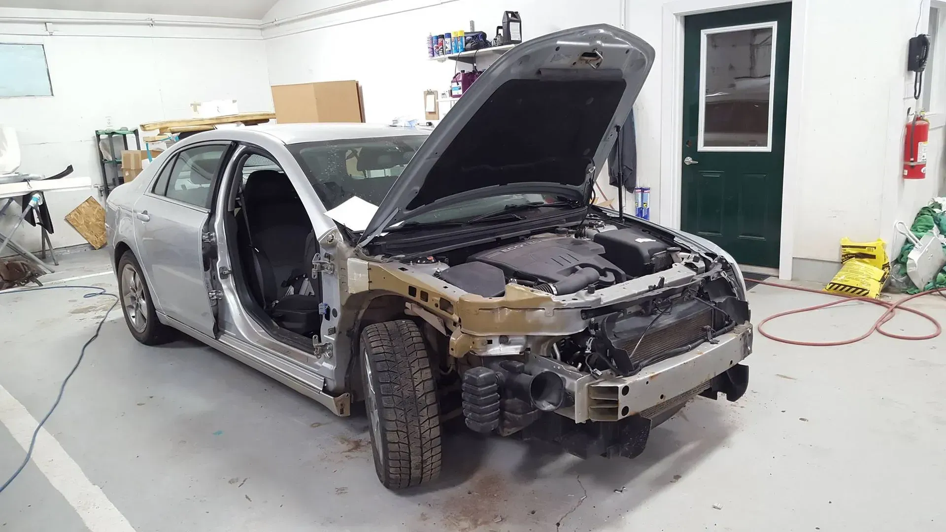 Silver car in auto body shop with front end disassembled and hood open on a repair stand