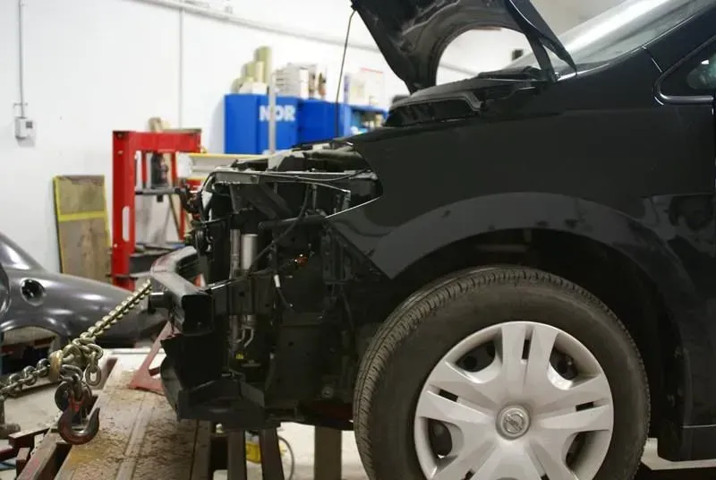 Black car with front end damage in an auto repair shop, hood open and bumper removed