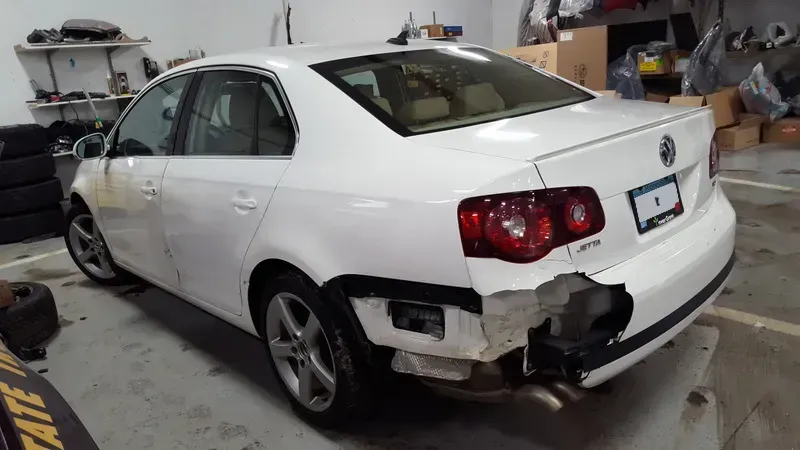 White sedan with rear-right body damage and missing bumper in a garage workshop.