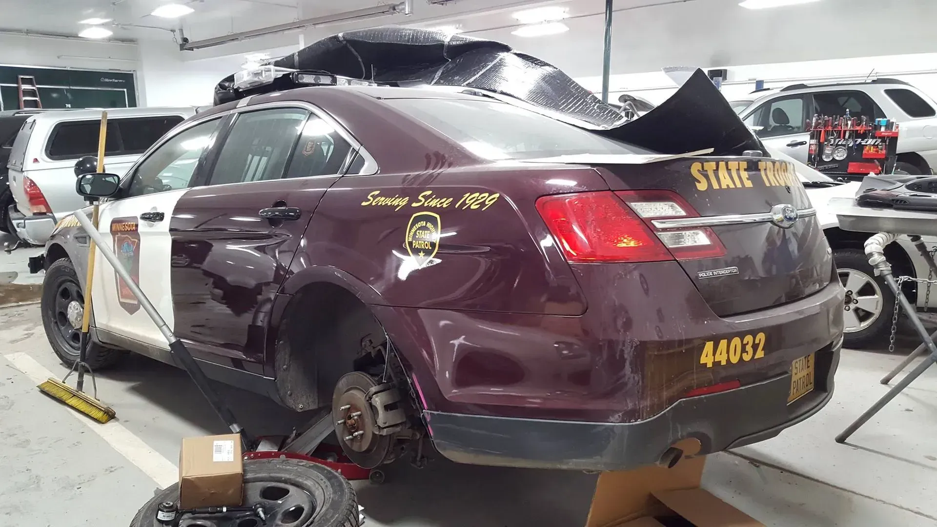Maroon SUV with missing rear wheel, parked in a repair garage with tools and other cars nearby