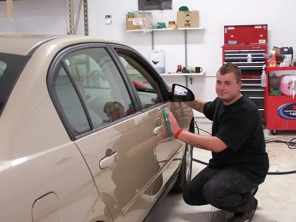 Man repairing a beige car door in a garage with tools and red tool cabinets.