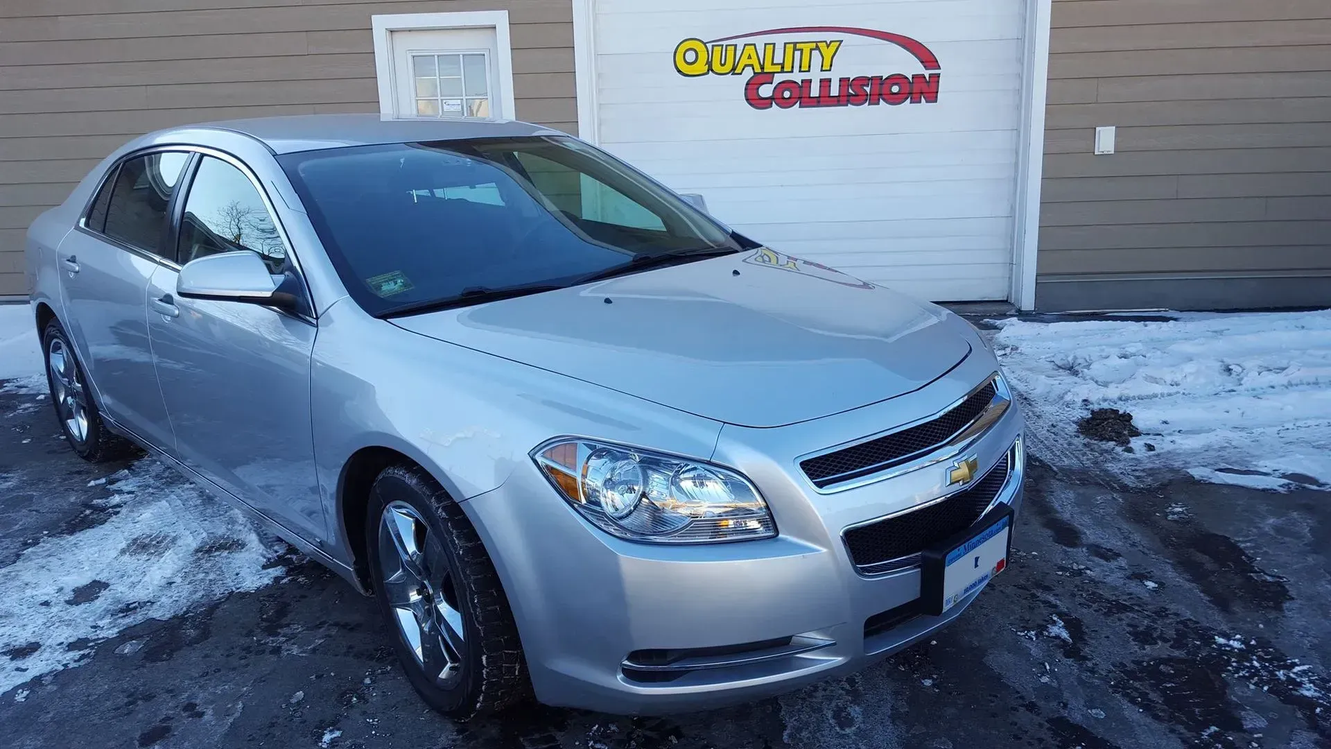 Silver sedan parked in snowy lot beside a building with a Quality Collision sign
