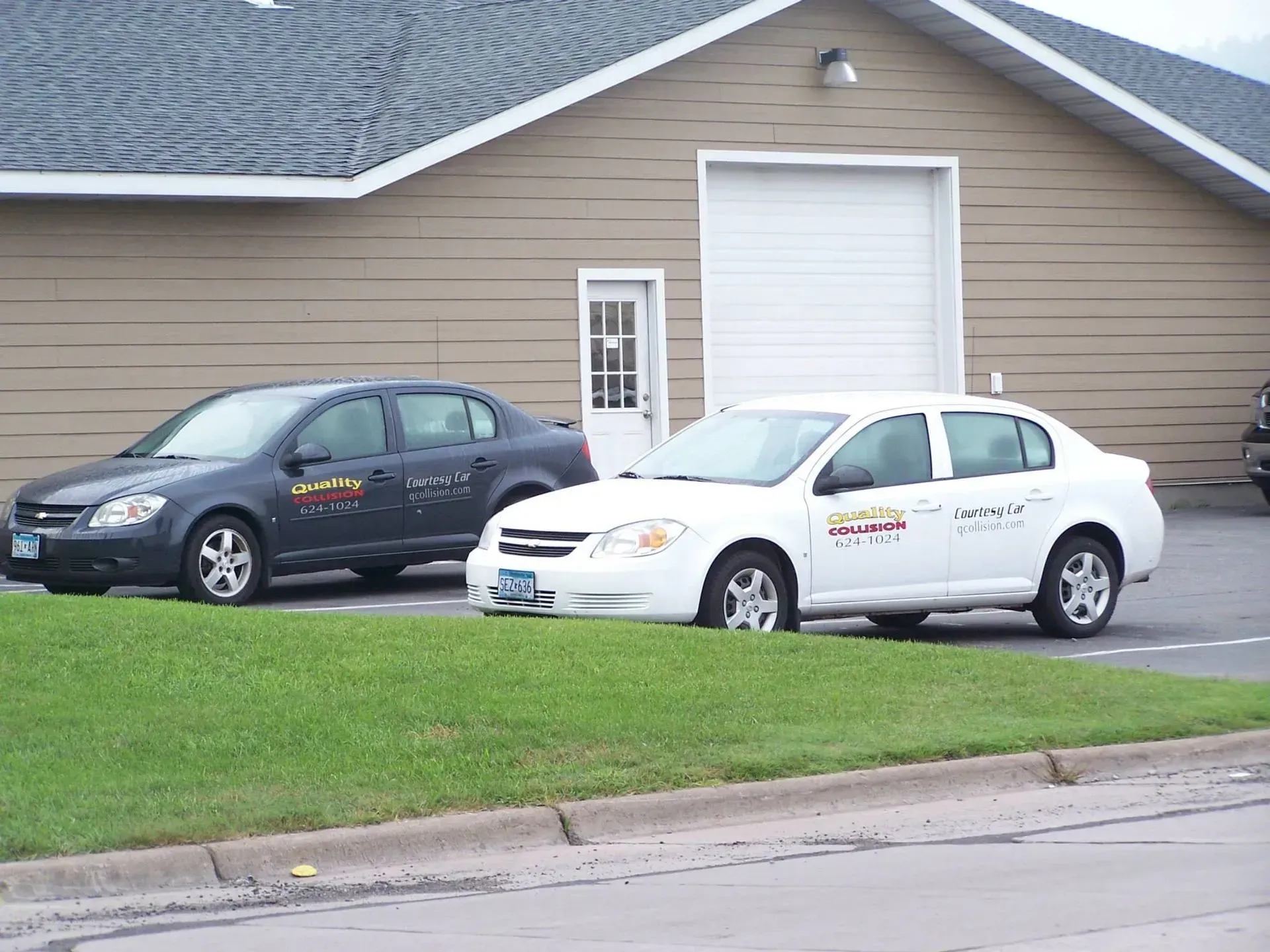 Two parked sedans in front of a beige garage, one black and one white, beside a green lawn.